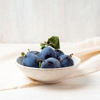 violet blue ripe plums in white bowl on white wooden table with linen textile ranner tablecloth