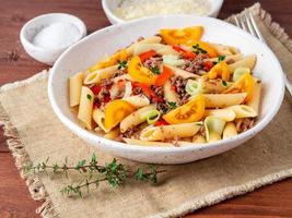 Penne pasta with yellow tomatoes, red and green vegetables, mincemeat on dark wooden background, side view