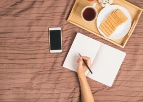 la mujer escribe en un gran cuaderno blanco abierto, se acuesta en la cama con un teléfono inteligente, bandeja de desayuno foto