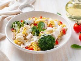 Pasta fusilli with vegetables, boiled steamed meat, white sauce on white wooden table, low-calorie food, low-fat diet, side view photo