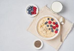Oatmeal with berries, chia, maple syrup and glass of milk on gray light background. Top view, copy space. Healthy diet breakfast