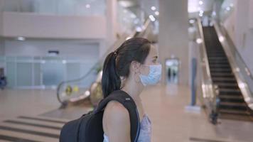 Aziatische jonge vrouw draagt een beschermend medisch masker dat met rugzak loopt op de roltrap van de luchthaventerminal, risico op reizen met het openbaar vervoer, nieuwe normale pandemie-uitbraak, covid19 corona-virus