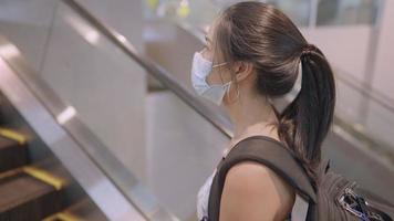 joven asiática que viaja sola durante la pandemia de covid19, usa una máscara protectora para prevenir enfermedades infecciosas, se para en una escalera mecánica con pasamanos, riesgo de objetos contaminados
