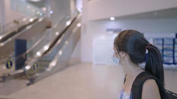 asiatisk ung kvinna bär ansiktsmask gå med ryggsäck inuti flygplatsterminalens rulltrappa i bakgrunden, avgång ankomst resehämtning, ombordstigningstid, kollektivtrafik, ny normal pandemi video