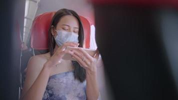 Asian woman wear protective face mask using smart phone while seated on window seat, inside aircraft cabin, relaxed comfortable new normal flight, public transportation risk of infectious diseases