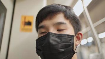 un homme asiatique portant un masque noir regarde la caméra puis regarde par la fenêtre, debout à côté de la porte du train. maladie pandémique covid-19, transports en commun, après une journée de travail fatigante, nouvelle heure de pointe de la vie normale video