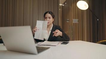 Young hardworking asian business woman doing video conference call presentation showing company analytics paper to the laptop computer screen, supervisor professional employee, late night meeting