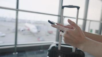les mains utilisant un smartphone se connectent en attendant au terminal de l'aéroport avant de monter dans l'avion, la réservation de billets de voyage, la valise de bagages du voyageur, l'aérobridge de la compagnie aérienne et les avions en arrière-plan