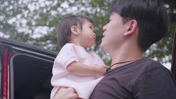 asiatico giovane adulto padre che tiene adorabile cute bambina figlia bambino piccolo in piedi accanto al veicolo sotto gli alberi all'aperto. bacio del bambino femminile asiatico sulla guancia di papà, legame di felicità del membro della famiglia video