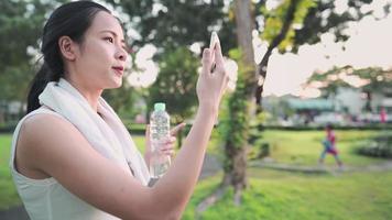 jeunes cheveux noirs asiatiques tenant une bouteille d'eau, faisant un selfie ou un appel vidéo, souriant et parlant en faisant du jogging dans le parc, distanciation sociale, verrouillage de la quarantaine covid-19, nouveau mode de vie normal