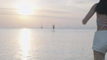 schuss einer jungen sexy asiatischen frau, die in den ozean läuft, genießen sie sonnenuntergangsstimmung, spritzendes wellenwasser, lustige, glückliche reiseferien am strand, tragen von bikini und shorts mit den beinen, die im wasser einweichen video