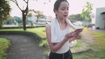 Aziatische vrouw die een handdoek gebruikt die zweet van het gezicht veegt, vrouw die smartphone schuift terwijl ze naar het park loopt tijdens het warme zonsonderganguur, ontspannen na de training, 5g draadloze technologie die mensen verbindt video