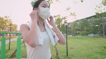 Young sporty white woman with headphones listening to music, smiling and taking off facial medical mask getting ready for outdoor exercise in public park. she holds surgical mask with hook on arm