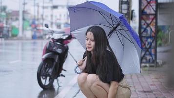 hübsches asiatisches mädchen, das regenschirm hält, setzt sich an regnerischen tag auf den straßenweg, regenzeit erreicht ihre hand, berührt regentropfen, bricht motorrad, fühlt sich enttäuscht im regen stecken video