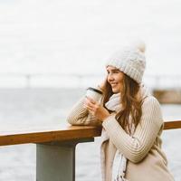 Beautiful young girl drinking coffee, tea from plastic mug in autumn, winter. A woman with long hair stands on waterfront on Baltic sea in port and waiting for ferry, heated by hot drink, copy space photo