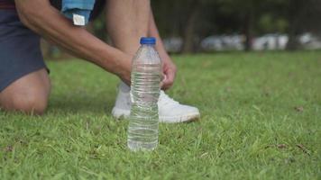Asian male runner tying shoe laces while exercising on green grass field, outdoor park, active lifestyle, stay hydrated water bottle placing on the side, active life insurance, morning workout routine