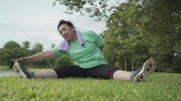 asiatisk aktiv överviktig mogen kvinna som gör kluvna ben sträcker hand till tå sitta på gräsmattan efter träning i utomhusparken, sjukförsäkring, flexibilitet i människokroppen, benmuskel full extension video