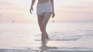 An unrecognized female legs in short white plant walks up from crystal clear ocean, with a view of huge mountain and sunset at skyline on background, asia natural recourses, summer tropical holiday, video