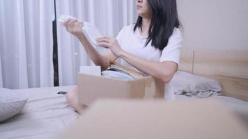 una joven asiática abre una caja de cartón de paquetes marrones, arroja cosas alrededor de la casa nueva, se sienta en la cama, una mujer soltera de vida soltera, se muda sola, se queda en casa con ropa informal