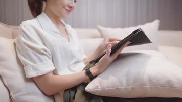 menina bonita asiática sentada no sofá confortável usando tablet digital dentro da sala de estar em casa, entretenimento de vida em casa, mídia social, feliz sorrindo e rindo assistindo conteúdo engraçado online video