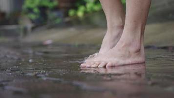 lage hoek van meisje met blote voeten springen en spelen op waterplassen op de regenachtige dag, stromende regen aan de straatkant wandelpad manier, leuke tijd opspattend water, goofy rondspringen, zorgeloze sfeer video