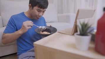 Asian young man enjoys watching a tablet while eating a spoon of riceberry in cozy living room, people and daily routine, having a meal, nutrition and delicious recipe, self entertainment at home video