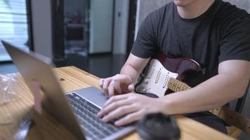 A young white skin man uploading his electric guitar recorded file into portable laptop on wooden office desk, human finger using computer touchpad, freelance guitarist preparing online performance video