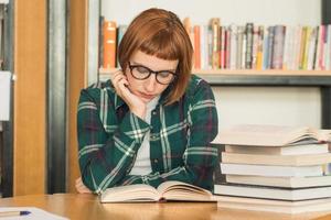 joven pelirroja con gafas lee un libro en la biblioteca foto