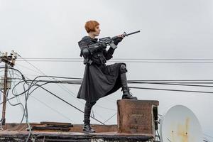 Young woman in modern black techwear style with rifle posing on the rooftop, portrait of redhead woman cyperpunk or post apocalyptic concept photo