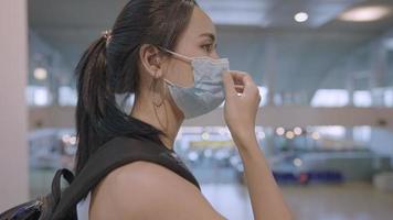 jovem mulher asiática tocando máscara facial protetora em pé na estação de metrô escada rolante, mochila de passageiros de viagem risco de doenças infecciosas edifício público, nova pandemia normal covid19 video