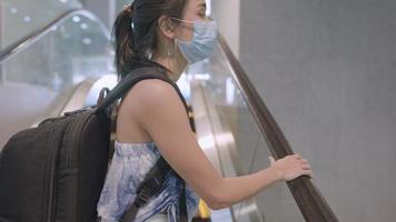 junge asiatische frau, die während der covid19-pandemie alleine reist, frau trägt eine gesichtsmaske, die vor infektionskrankheiten schützt, geht von der rolltreppe im seitlichen flughafenterminal hoch, reiserisiko, soziale distanz video