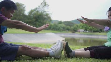 asiatische Seniorenpaare strecken ihre Beine von Hand bis Fuß, während sie sich während des Trainings im öffentlichen Park im Freien auf das Gras setzen, gesundes Ruhestandsleben im Alter, Familienzeit, Seeberghintergrund video
