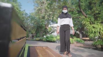 Asian Muslim woman wear hijab and face mask. Taking a break sit down on the park bench under trees on sunny day, using smartphone, New normal COVID pandemic distancing network connection online video