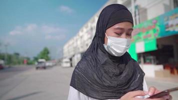 asiatische junge muslimische Hijab-Frauen tragen eine Gesichtsmaske. frau, die smartphone benutzt, während sie auf der städtischen straßenseite geht, auto, das die hintergrundnetzwerkverbindung weitergibt, covid-19-pandemie neues normales, modernes leben