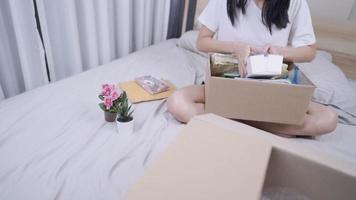 mujer joven asiática sentada dentro del dormitorio, mudándose a un nuevo lugar con una caja de cartón en su regazo, sacando una foto antigua de la caja a un nuevo apartamento, clasificando la limpieza, reubicando la memoria en movimiento
