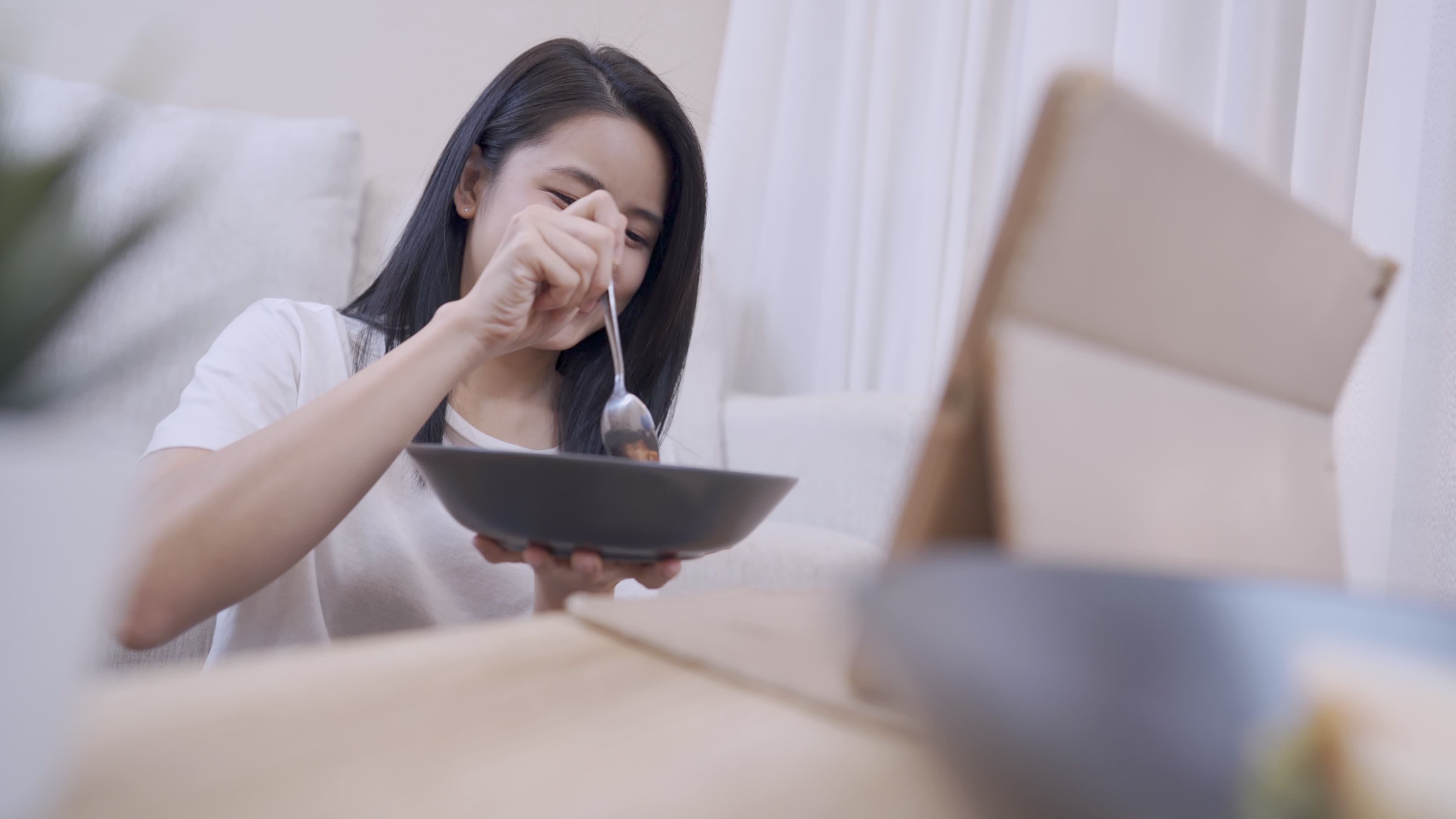 Young Asian woman eating her dinner while watching online content fun laughing, sit down on the floor with couch on the back inside apartment living room, simple living wellness, stay at home alone