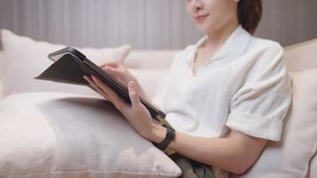 femme asiatique relaxante assise sur un canapé confortable à l'aide d'une tablette coulissante à la main dans le salon de la maison, divertissement de la vie à la maison, travail à domicile, liberté de travail, designer indépendante travaillant sur un canapé video