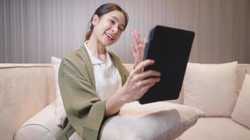 Aziatische vrolijke freelance ontwerper met een betrokken videogesprek via tablet in haar werkproces terwijl ze in een gezellige huiskamer zit, digitale online conferentie, communicatieconcept op afstand video