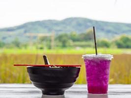 Noodle soup in black cup with red chopsticks and herb water on white wooden table. photo