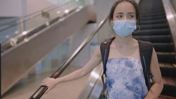 Asian Young female traveler in  protective face mask walking out from escalator with backpack inside train station, corona virus prevention outside public places, new normal pandemic awareness video