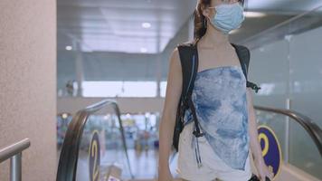 une jeune femme asiatique porte un masque protecteur en sortant de l'escalator avec un sac à dos à l'intérieur d'un terminal d'aéroport vide, risque de maladies infectieuses dans les lieux publics, nouvelle pandémie normale, assurance voyage