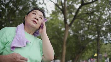 Aziatische senior vrouw veegt zweet van haar gezicht terwijl ze rust tijdens het sporten in het park, buiten staan en rusten na het sporten, pensioen leven, gezondheidstoestand risico op borstkanker bij vrouw video