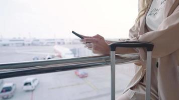 cerrar las manos de las mujeres de negocios usan mensajes de texto de teléfonos móviles mientras esperan el vuelo, se paran dentro del viaje de negocios de la terminal del aeropuerto, el teléfono celular y el equipaje de viaje y el avión en el fondo video