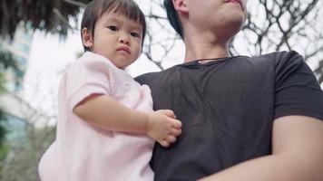 young Asian father holding his baby girl standing inside park under trees, child care parenting bonding, pure oxygen fresh air, children innocence, father and daughter looking at camera, small child video