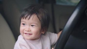 Asiatisches süßes Kleinkind, das Spaß beim Spielen im Auto hat und mit dem Lenkrad spielt, Baby, das auf dem Autofahrersitz sitzt, glückliches kleines Mädchen, das sich selbst genießt, indem es vorgibt zu fahren, Vorschulenergie