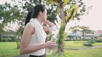 Young beautiful athletic woman directly drinks mineral from reusable bottle with wipes sweat using white towel from her face and neck after jogging in green beautiful park, feeling fresh and relax video