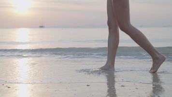 Low angle young woman legs bare foot walking along sunset wavy sand beach enjoy golden sunset in distance feeling nature with the horizon seascape and sky, tropical paradise travel destination video