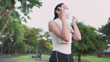 jonge fitte witte aziatische arbeider die sportkleding draagt, doet een medisch masker af en zet een koptelefoon op, geniet van muziek luisteren en sporten vroeg op een zonnige dag in het groene stadspark, gezond stadsleven
