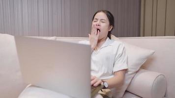 Pretty asain woman yawning while get online with laptop computer sitting on comfortable sofa, work exhaustion late at night, sleepy tiring working woman inside home workspace, not enough rest video