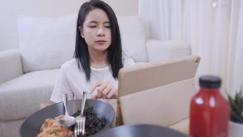 junge asiatische frau, die mit löffel und gabel isst, während sie sich im wohnzimmer amüsiert, sich mit couch dahinter hinsetzt, einfacher lebensstil, allein zu hause bleibt, positive weibliche, gesunde hausmannskochabendessen video